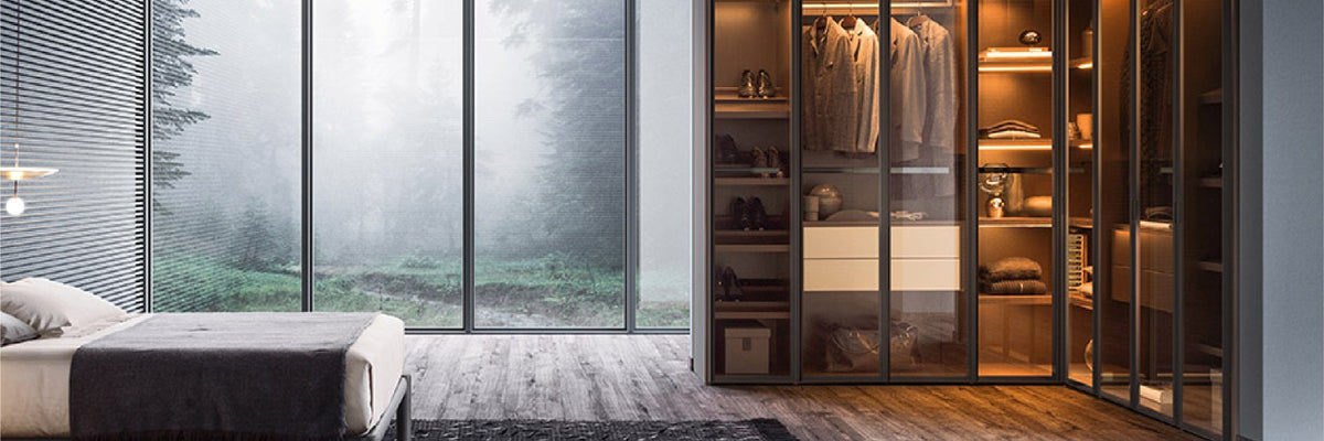 Milano glass-front walk-in closet system, lit from inside, showing hanging clothes
