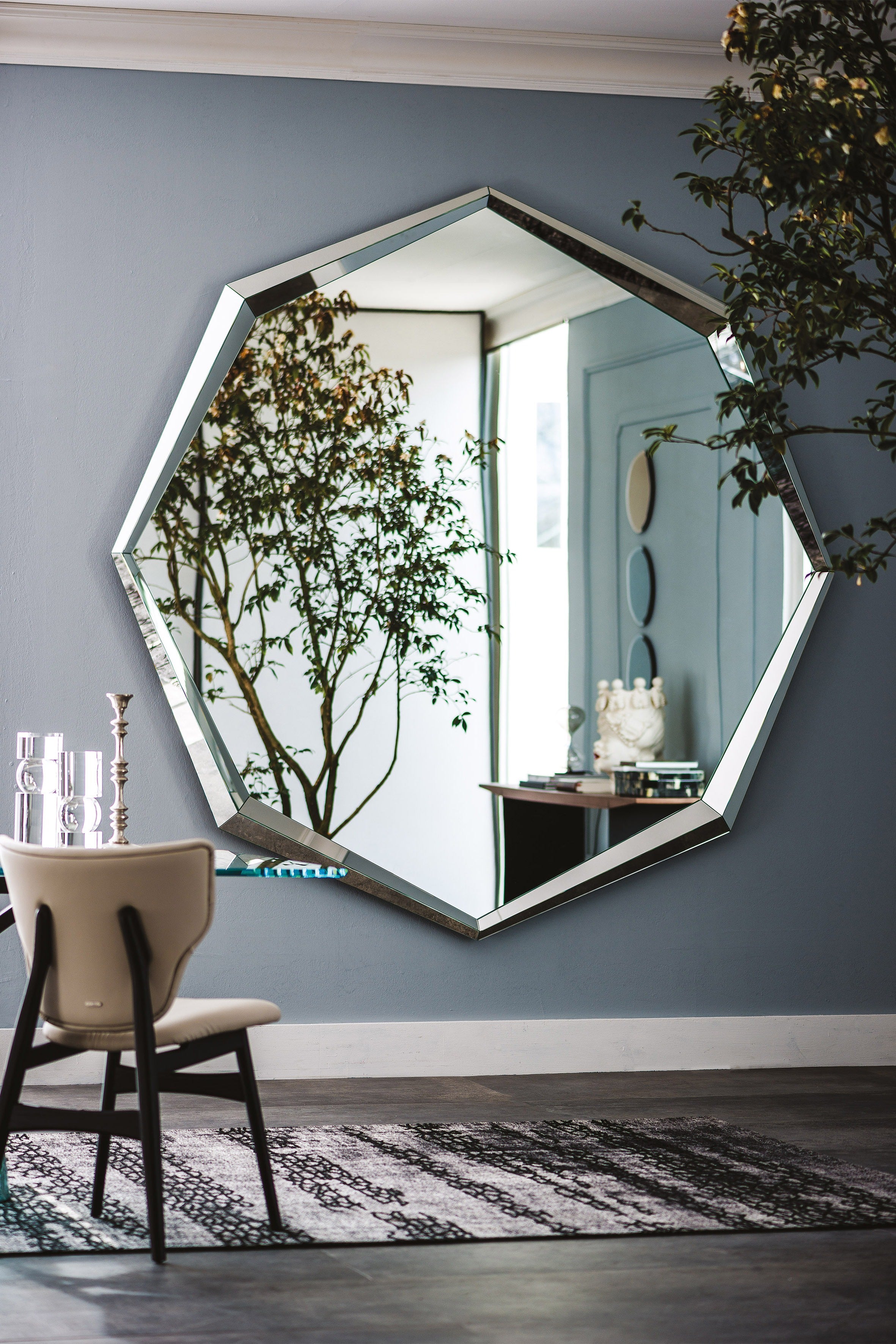 Emerald Magnum mirror on a blue-grey wall with a cream upholstered chair, a rug and a small tree