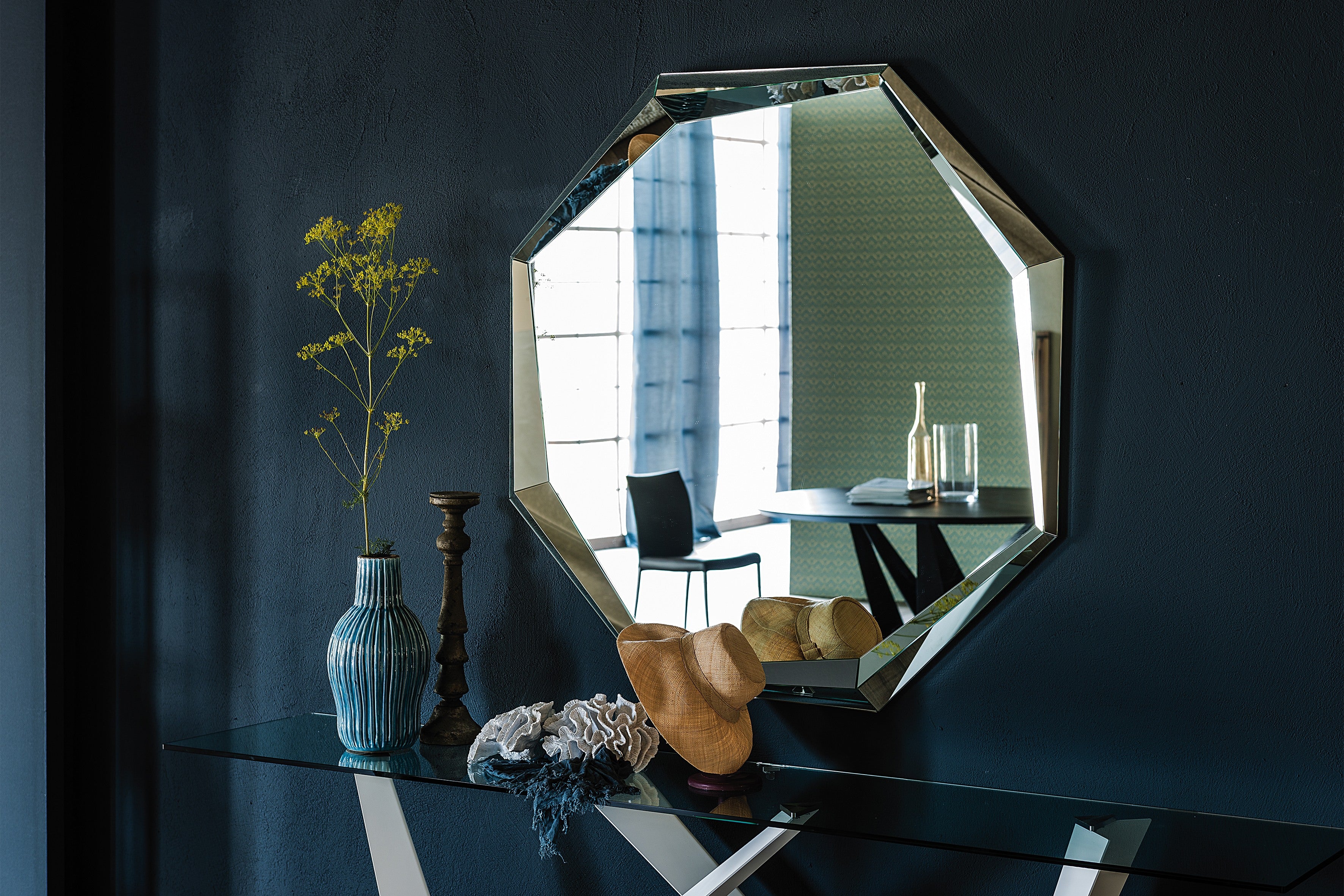 Octagonal Emerald mirror in clear glass on a dark blue wall above a glass console with a vase
