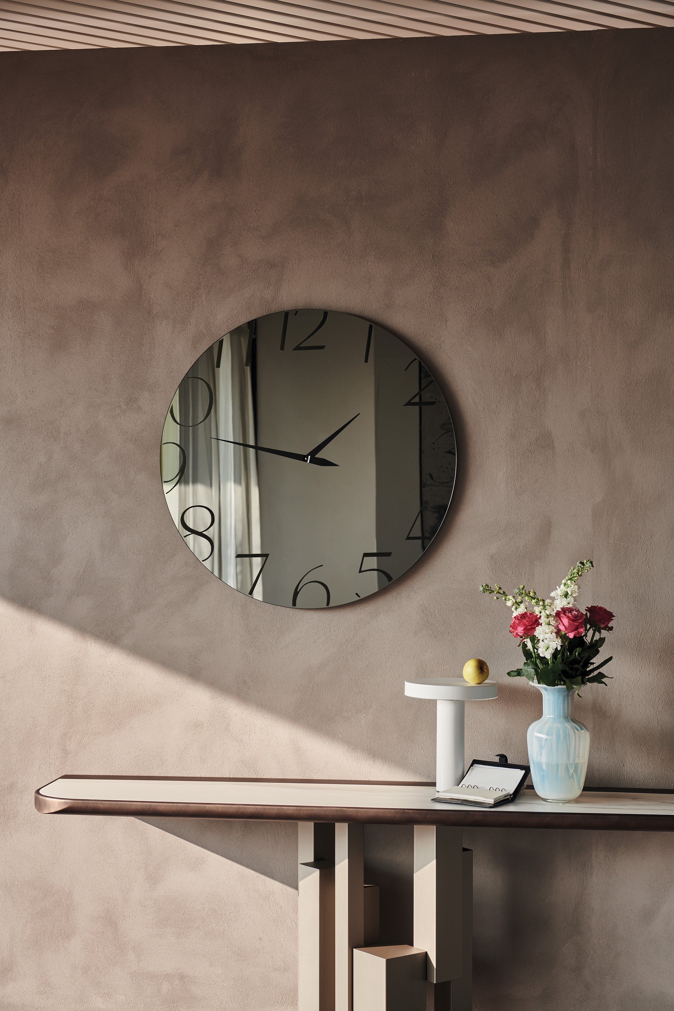 Moment mirror: round fumé Moment clock-mirror on a taupe plaster wall above a slim console with a lamp and a vase of roses