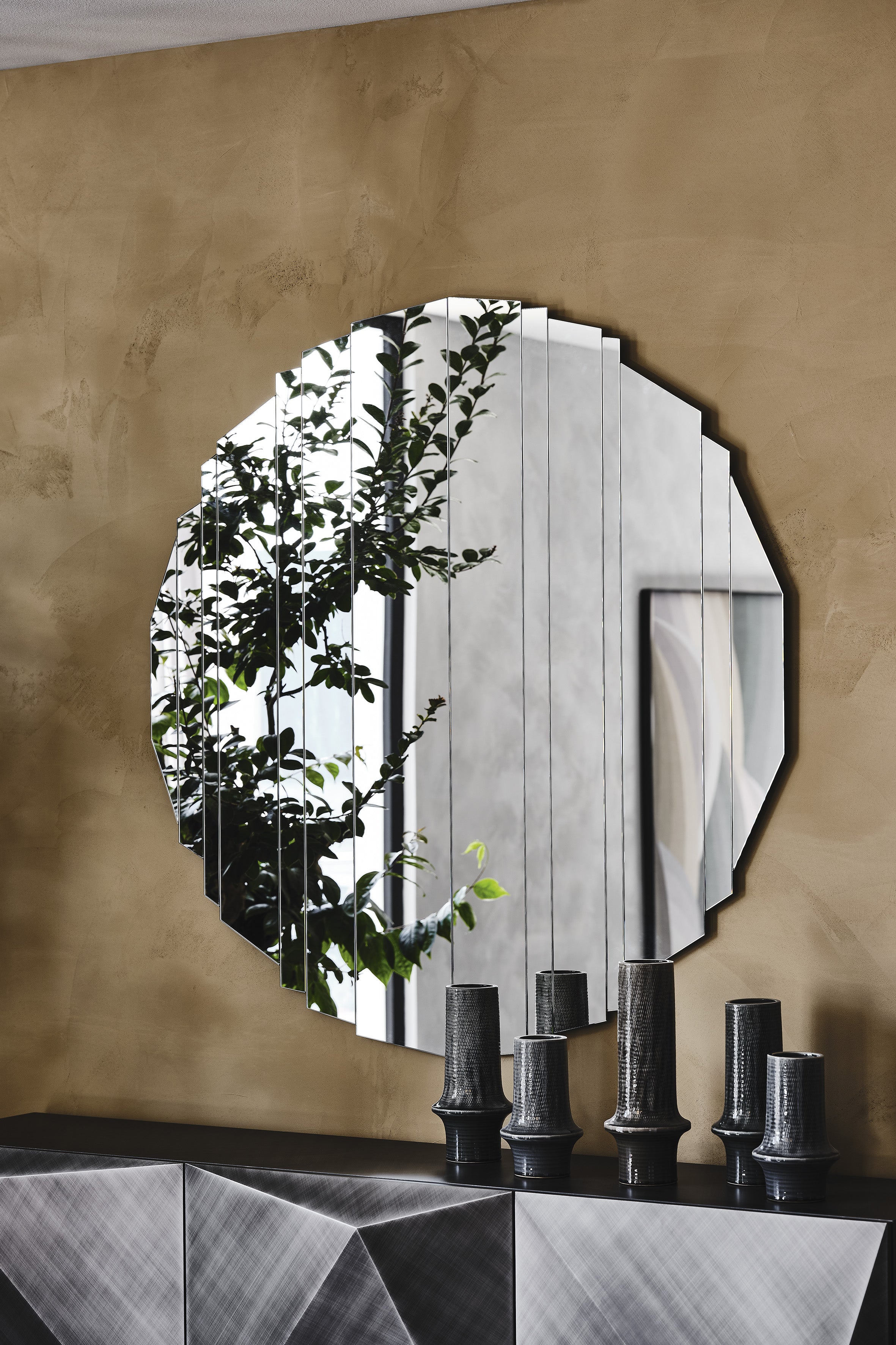 Stripes mirror: round Stripes on a warm gold-plaster wall over a faceted sideboard with dark vases, reflecting a plant