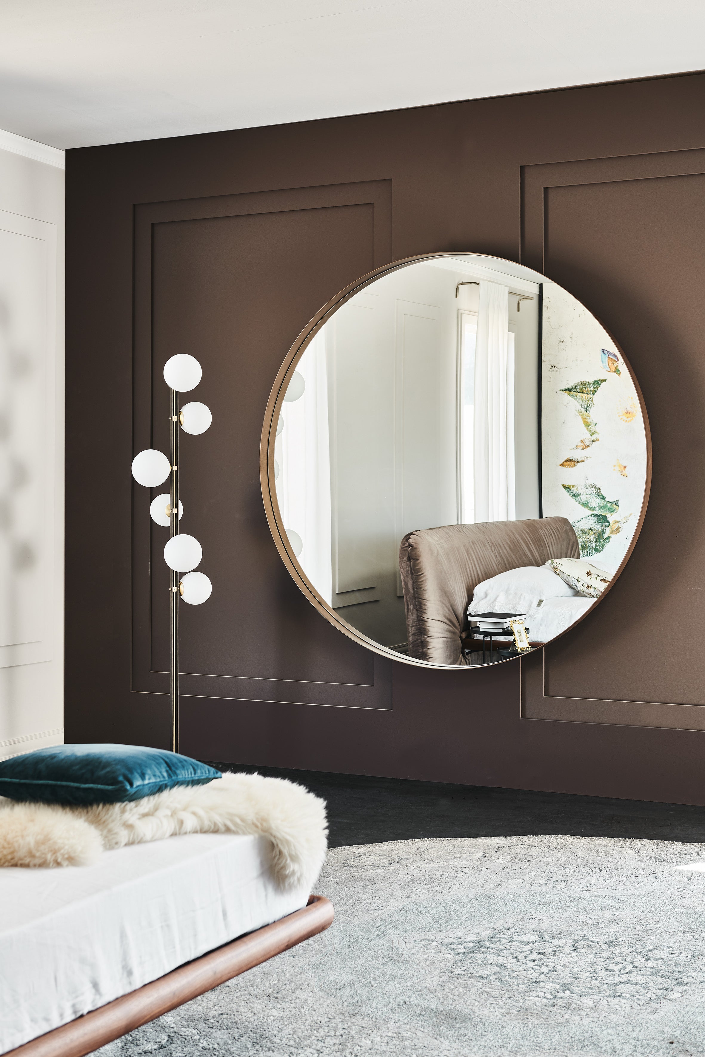 Round bronze-framed Wish Magnum mirror on a brown panelled bedroom wall with a globe floor lamp